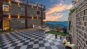 Une terrasse extérieure à carreaux avec un espace de jeux comprenant un toboggan et des jouets, et une vue sur les montagnes en arrière-plan.