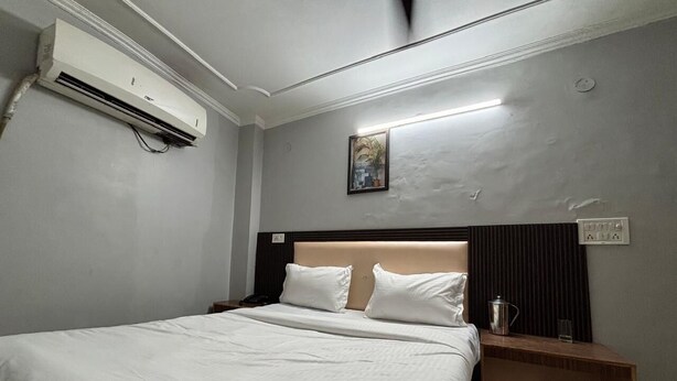 A hotel room with a bed, bedside tables, a nightstand, an air conditioning unit, a wall-mounted light, and a painting. - The Capstone (New Delhi)