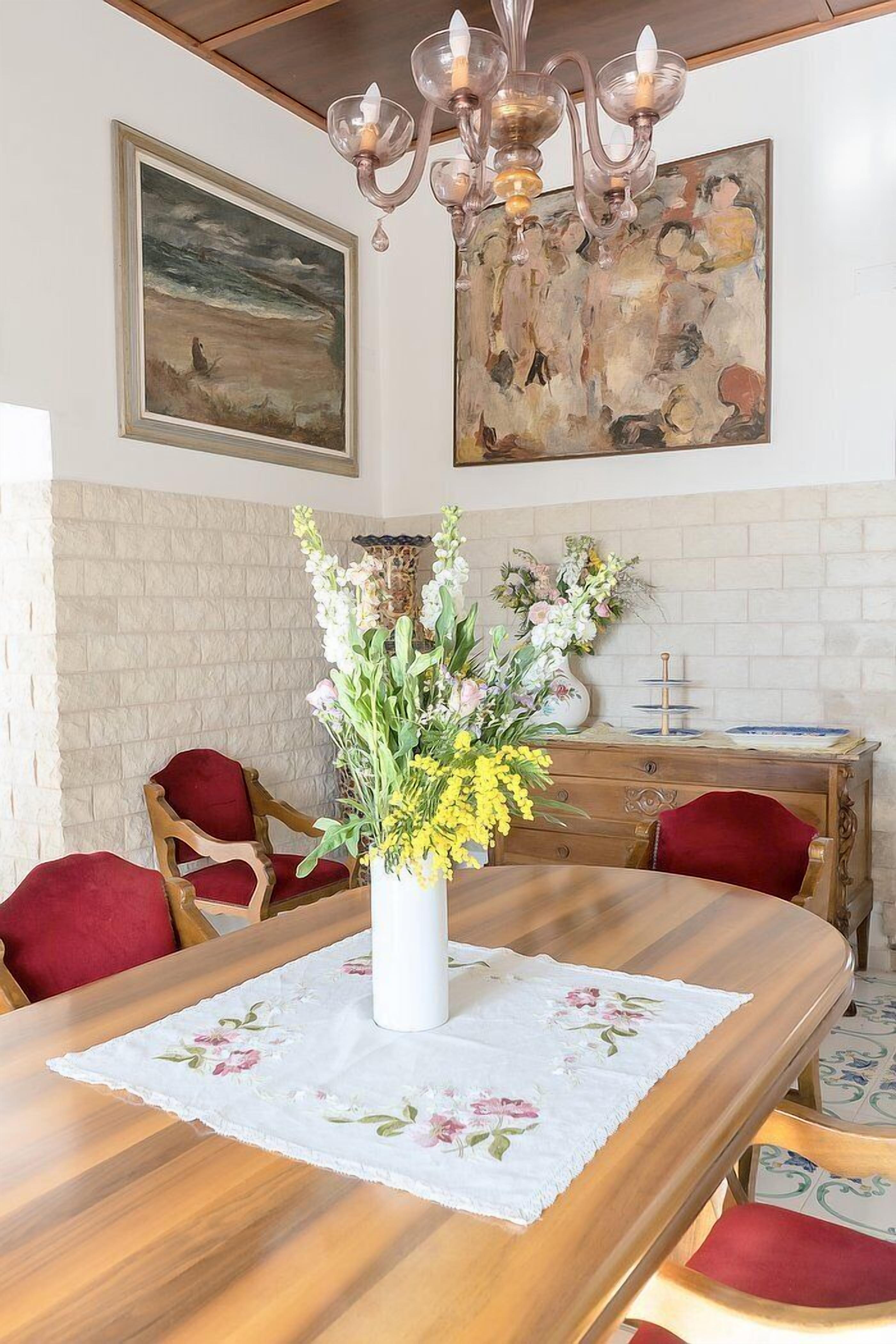 Un coin repas avec une table en bois, des chaises à revêtement rouge, un vase blanc orné de fleurs et un lustre.