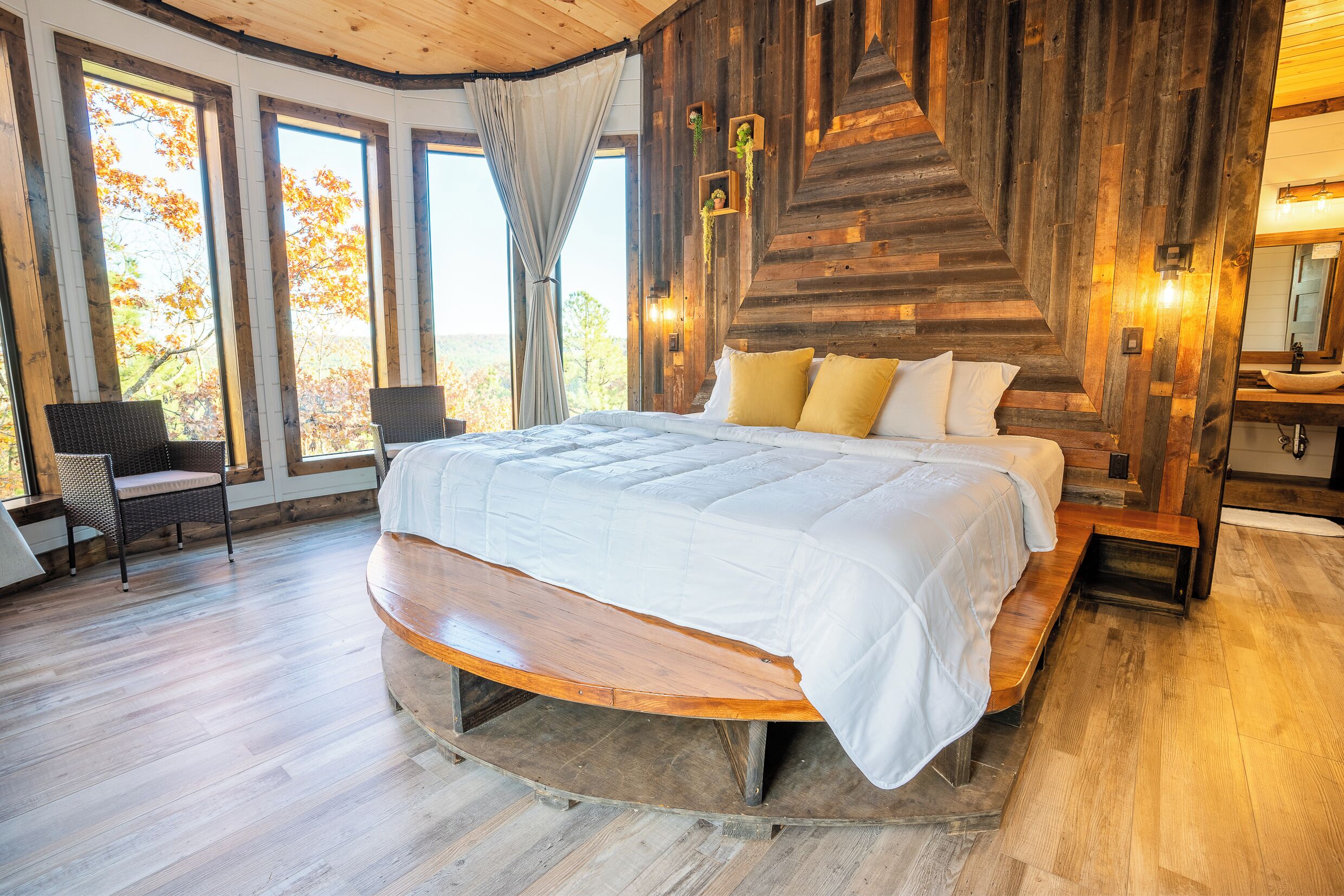 Una cama grande con un banco de madera, una silla y vistas a árboles otoñales a través de amplias ventanas.