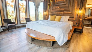 A large bed with a wooden bench, a chair, and a view of autumn trees through large windows.
