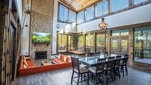 A spacious dining area with a large wooden table and chairs, a stone fireplace, and a view of the outdoors through large windows.