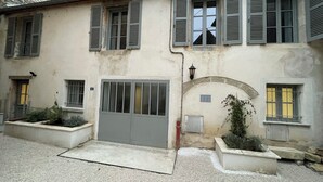 Un edificio de dos pisos con una puerta gris, varios ventanales con contraventanas y un pequeño jardín con plantas.