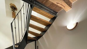 Una escalera de caracol con escalones de madera y barandilla de metal, un barril antiguo sobre un taburete de madera, y una pequeña caja de madera en el suelo.