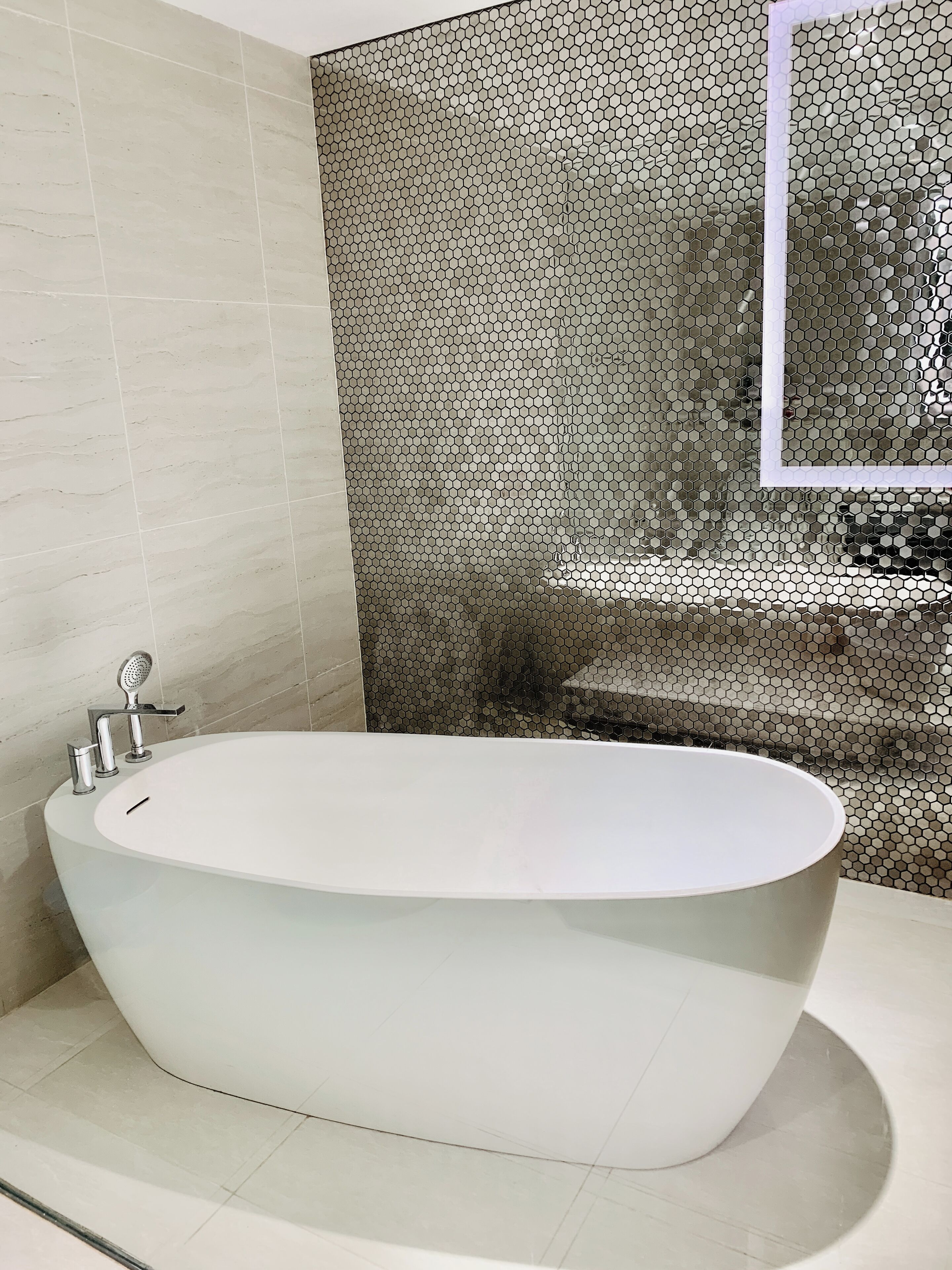 A modern bathroom with a freestanding bathtub, a shower with a handheld showerhead, and a tiled wall with a honeycomb pattern.