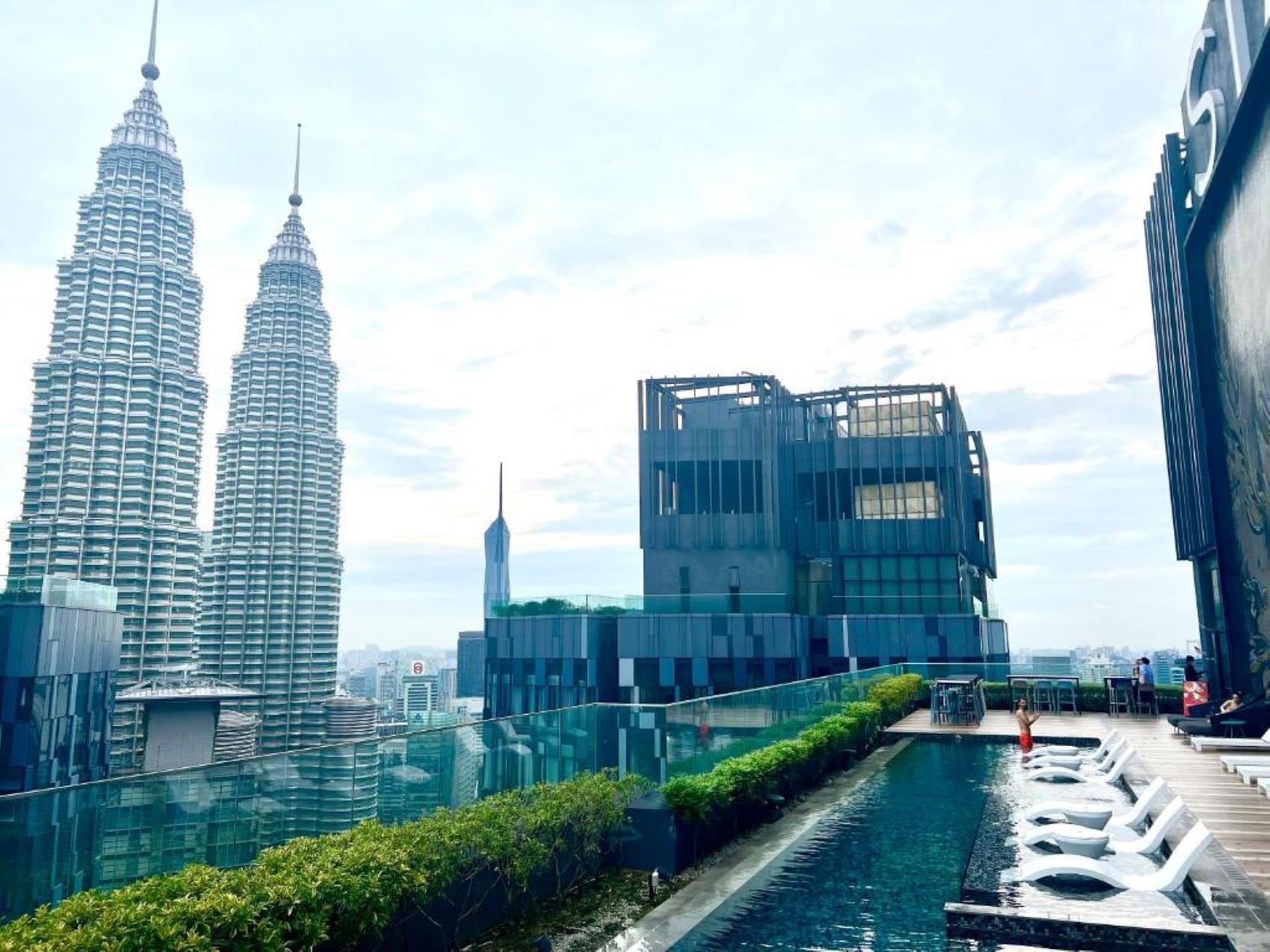 A modern building with a glass facade, a rooftop pool with lounge chairs, and views of the Petronas Towers.