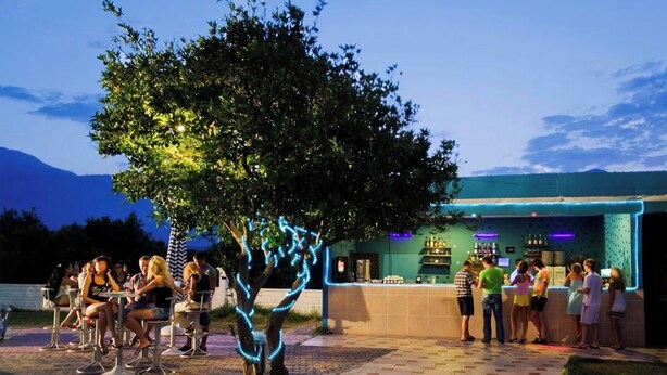 An outdoor seating area with tables and chairs, a tree with illuminated lights, and a bar area with people standing and conversing.