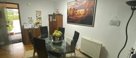 A dining area with a glass table, black chairs, a flower centerpiece, a cityscape painting, a wooden cabinet, and a radiator.