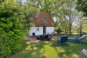 Een klein huis met pannendak, een terras met een tafel en stoelen, en een gazon.