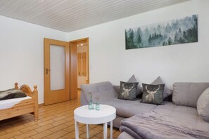 Ein Zimmer mit einem Bett, einer Couch, einem runden Tisch mit Vasen, einer Tür und einem Wandbild mit Waldmotiv.