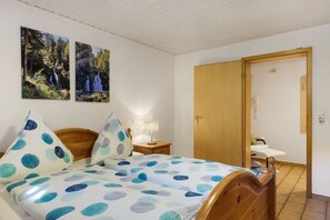 Ein Schlafzimmer mit einem Bett, zwei an der Wand befestigten Bildern, einem Nachttisch mit Lampe und einer Holztür, die in einen anderen Raum führt.