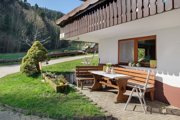Un ensemble de banc et de table en bois est installé à l’extérieur, avec un bâtiment blanc en arrière-plan.