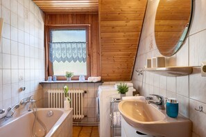 A bathroom with a bathtub, sink, and toilet, wooden ceiling, and a window with a curtain.
