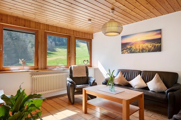A living room with a wooden ceiling, a black leather sofa, a wooden coffee table, and a large window with a view of a green landscape.