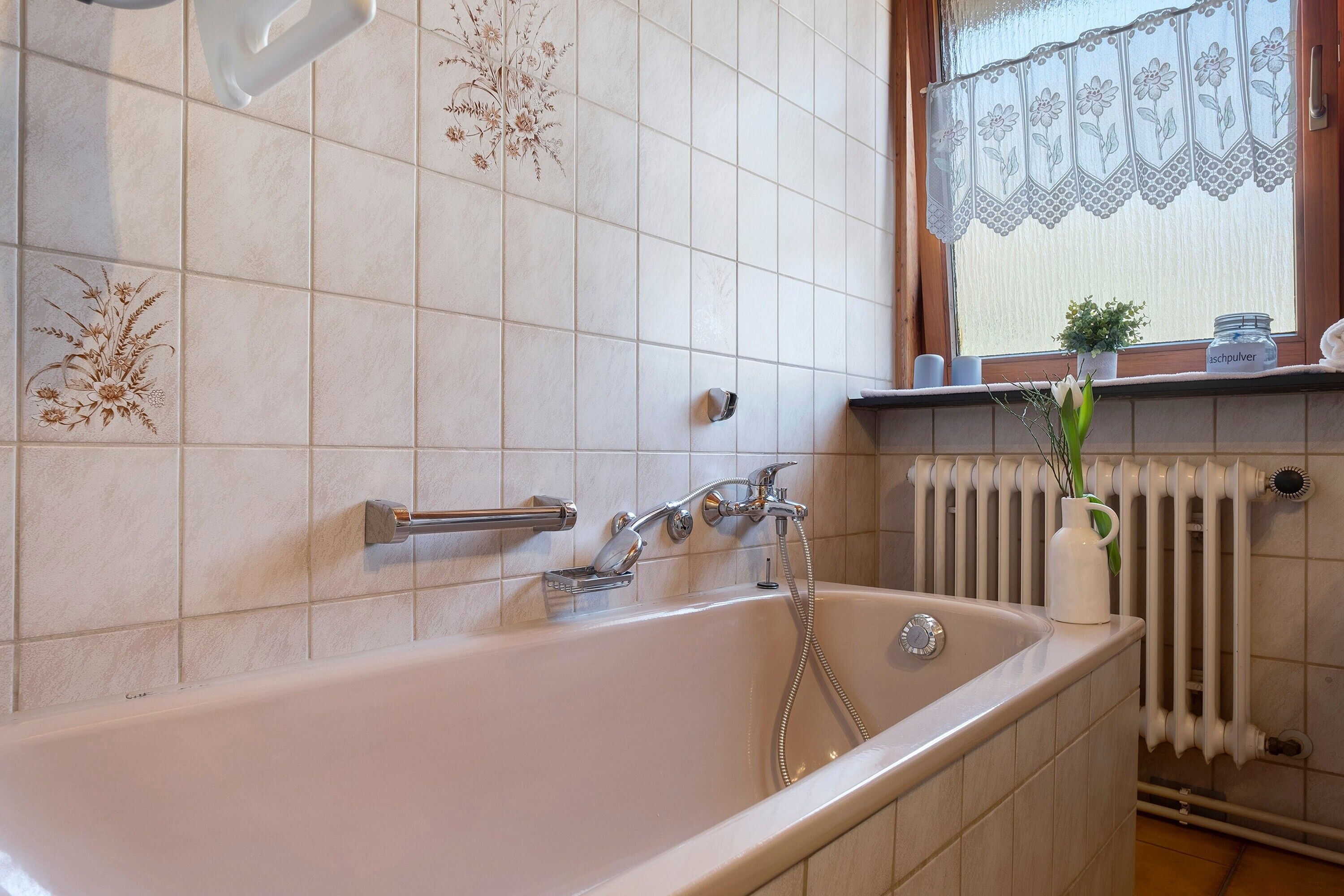 A bathroom with a bathtub, a window with lace curtains, a radiator, and a shelf with a plant and a jar.
