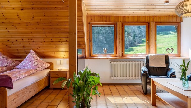 A wooden interior with a bed, a chair, a table, and a window with a view of greenery. - Apartment Lavendel with Mountain View, Shared Terrace and No Wi-Fi (Kirchzarten)