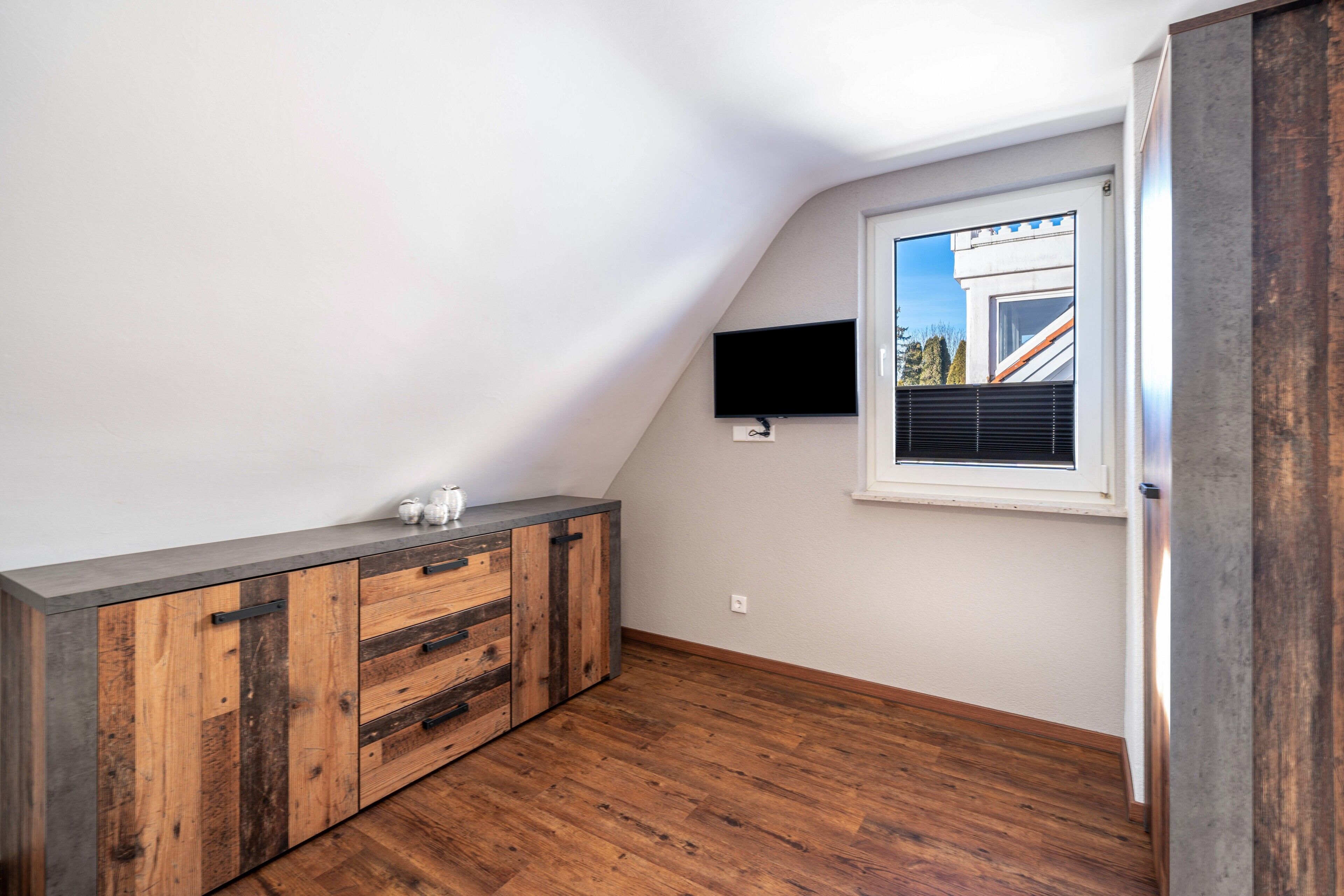 Una habitación con techo inclinado, un armario de madera empotrado, un televisor de pantalla plana en la pared y una ventana con persianas.