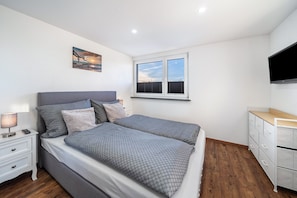 Un dormitorio con cama, mesita de noche, lámpara, ventana y televisión montada en la pared.