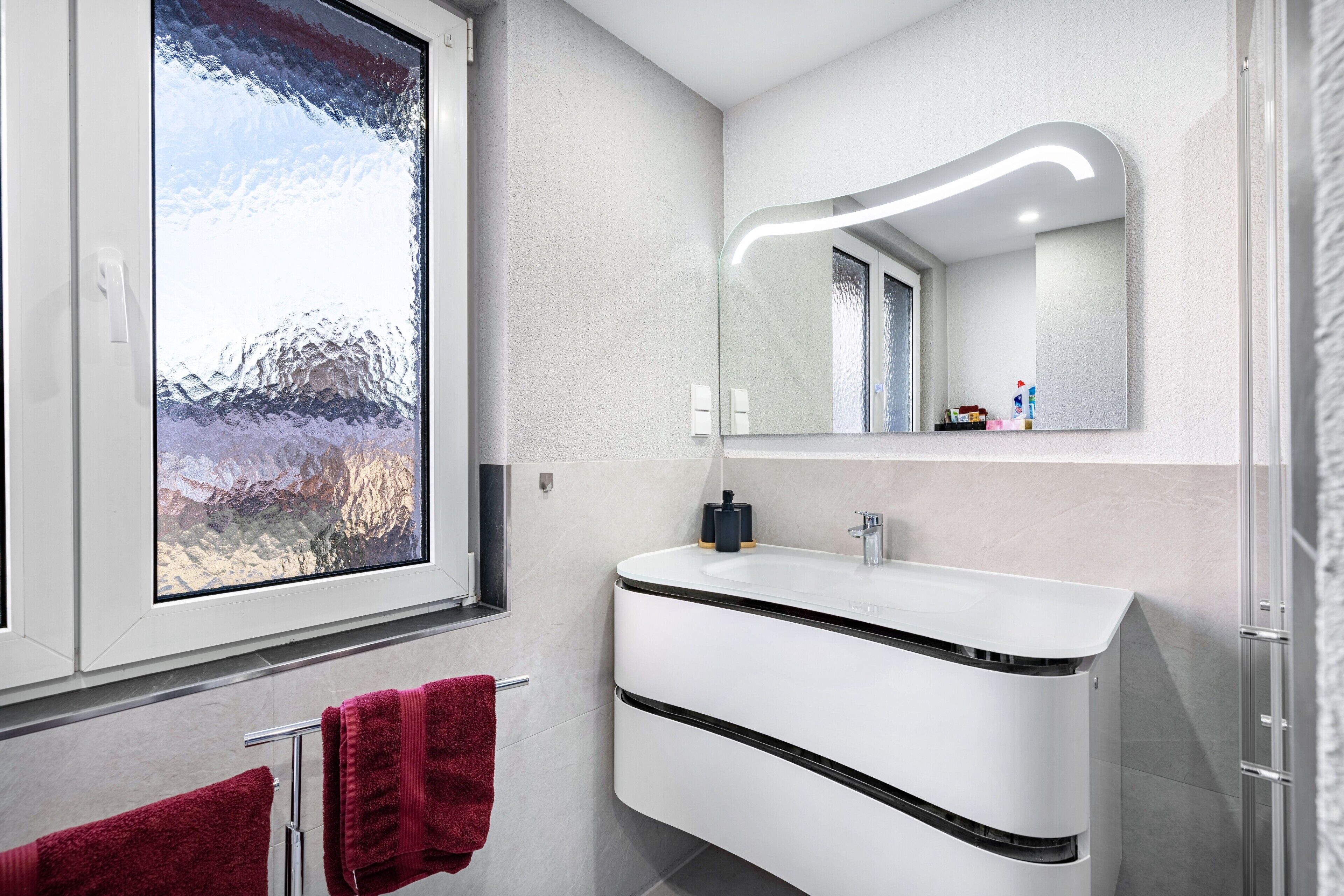 A modern bathroom with a large window, a white sink, and a mirror with built-in lighting.