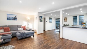 Una sala de estar moderna con un sofá gris, suelo de madera y una zona de cocina con encimera blanca y persianas azules.
