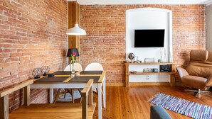 Een woonkamer met een bakstenen muur, een flatscreen televisie, een houten tafel, een bruine leren fauteuil en een kleurrijk vloerkleed.
