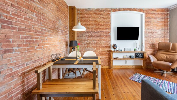 Een kamer met een bakstenen muur, houten vloer, een eettafel met stoelen, een bank, een aan de muur bevestigde televisie en een hanglamp.