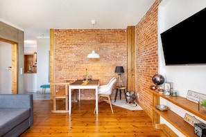 Ein modernes Wohnzimmer mit Backsteinwand, Holzboden, einem Esstisch mit Stühlen, einer Couch, einem an der Wand befestigten Fernseher und einem kleinen Regal mit Dekoration.