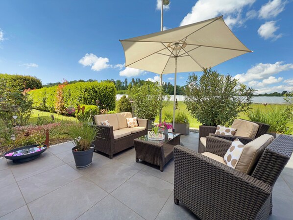 An outdoor patio area with wicker furniture, a large umbrella, and a view of a landscaped garden.