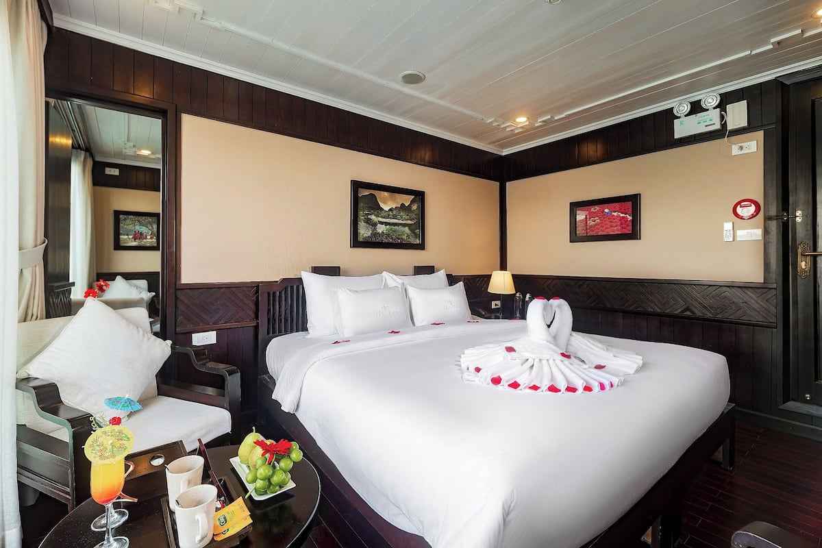 A hotel room with a large bed, a sitting area with a chair, a small table with a drink and fruit, and two framed pictures on the walls.