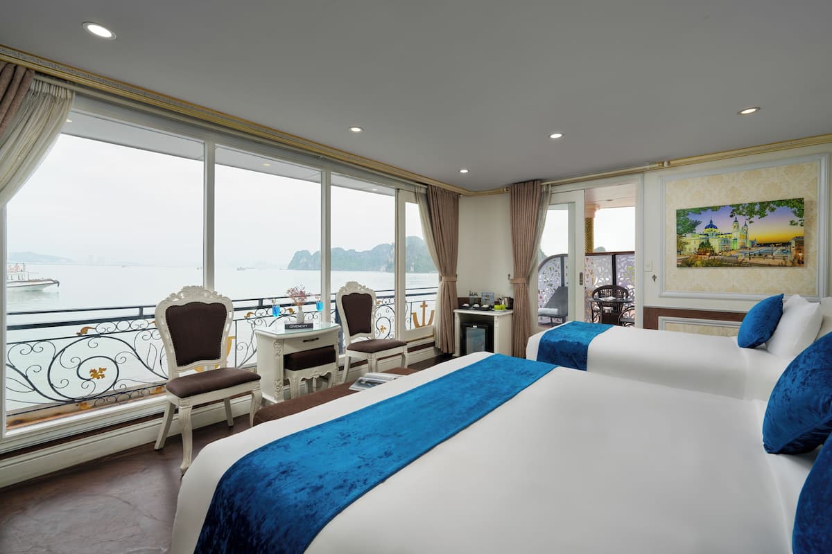 A hotel room with two beds, a desk, and a chair by the window with a view of the sea.