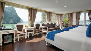 A hotel room with a large bed, a desk with a chair, a small table, and a view of a mountainous coastline. - Halong Aquarmarine Royal Cruise  (Tuan Chau)