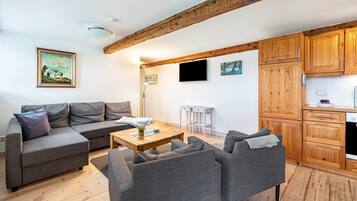 En moderne stue med sofa, lænestole, sofabord og TV. Der er en synlig træbjælke i loftet, et billede på væggen og et køkkenområde med træskabe.