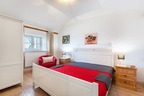Une chambre à coucher comprenant un lit, un fauteuil rouge, une table de chevet, une fenêtre avec des rideaux et deux œuvres d’art accrochées au mur.