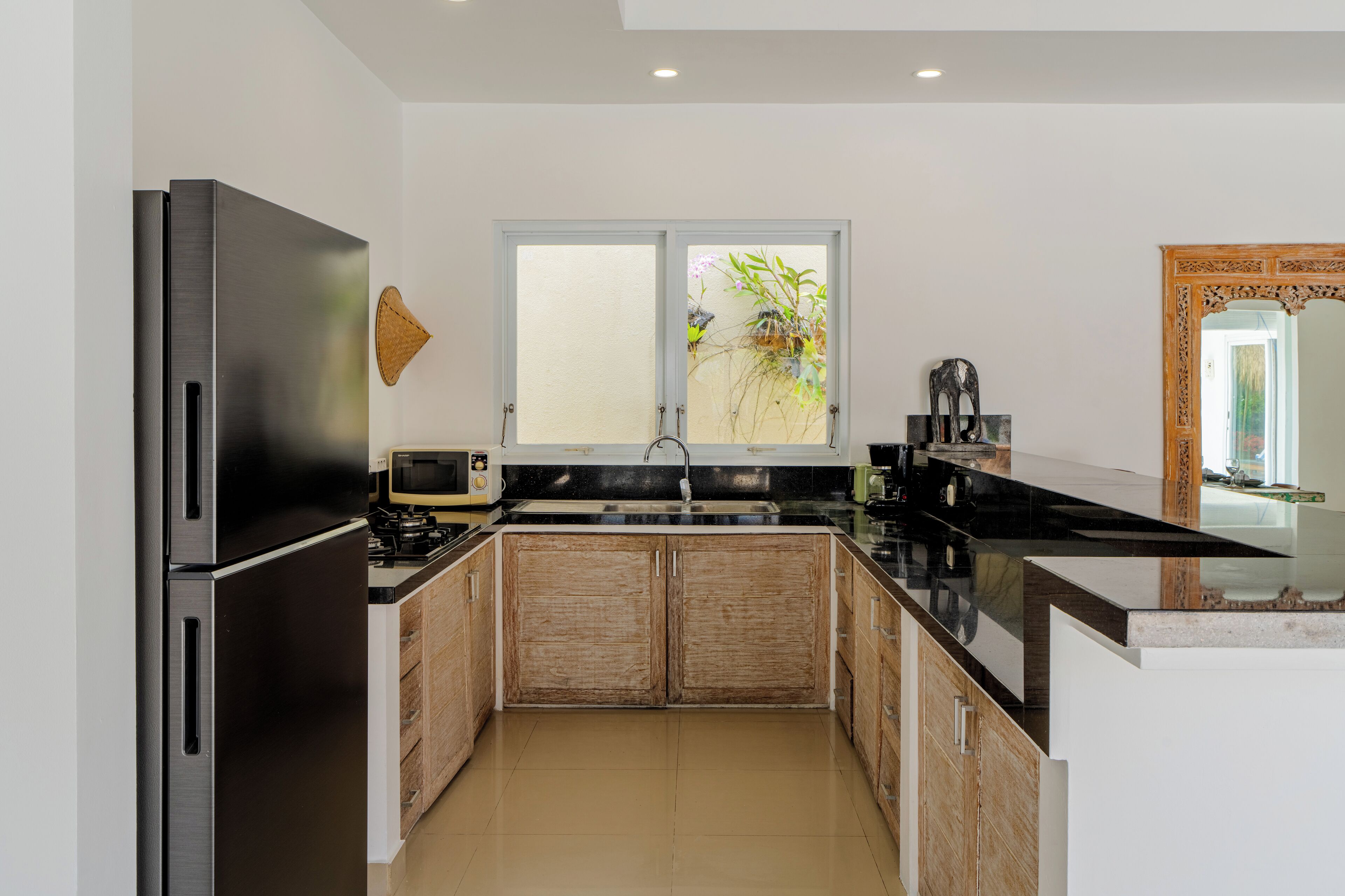 A modern kitchen with a stainless steel refrigerator, wooden cabinets, a black countertop, and a window with a view of greenery.