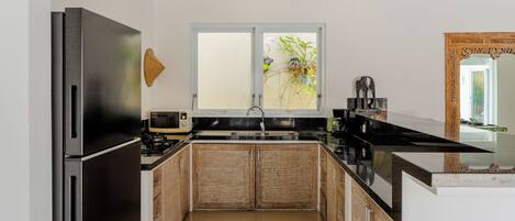 A modern kitchen with a stainless steel refrigerator, wooden cabinets, a black countertop, and a window with a view of greenery.