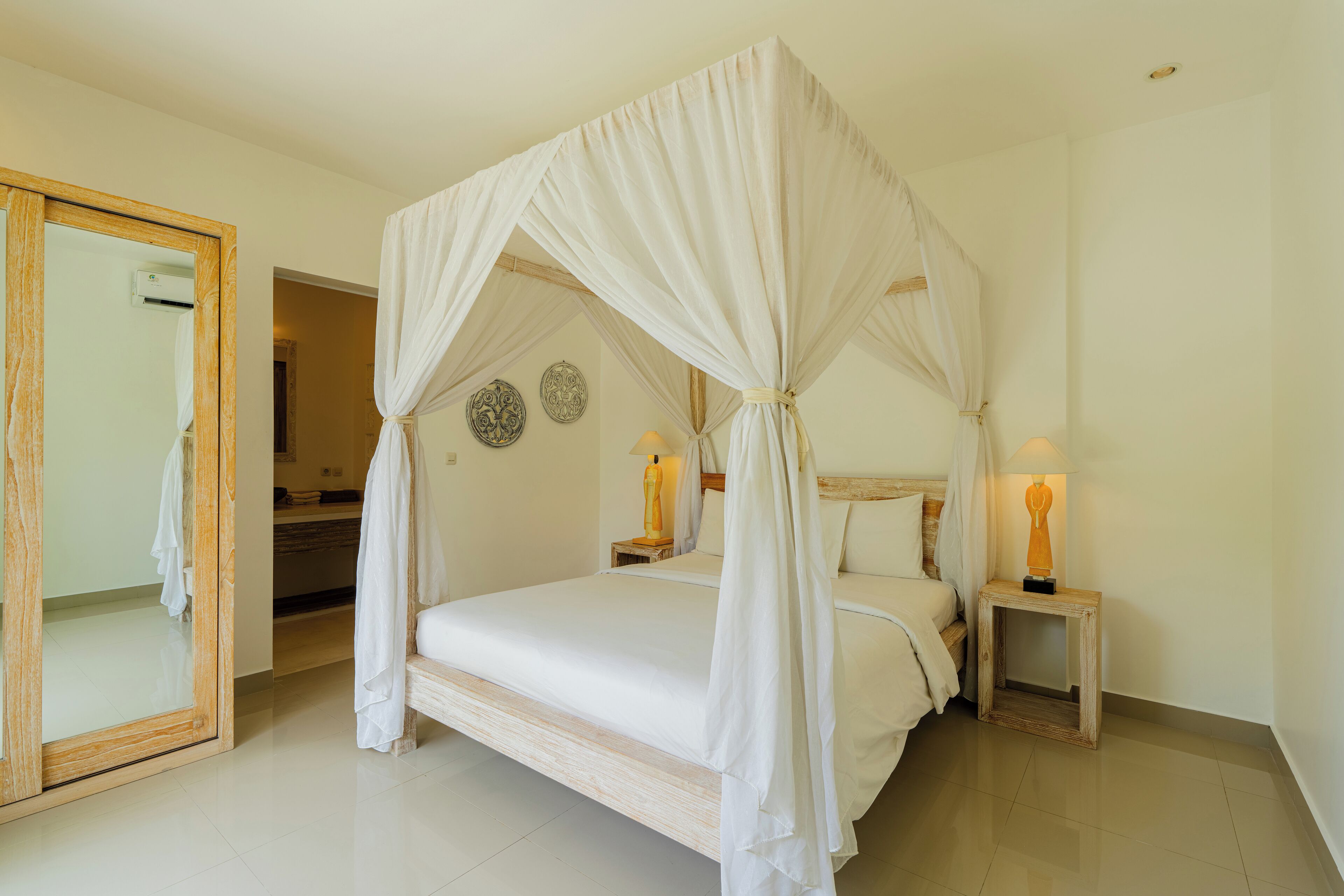 A four-poster canopy bed with white bedding, two bedside tables with lamps, a wooden-framed mirror, and decorative wall plates.