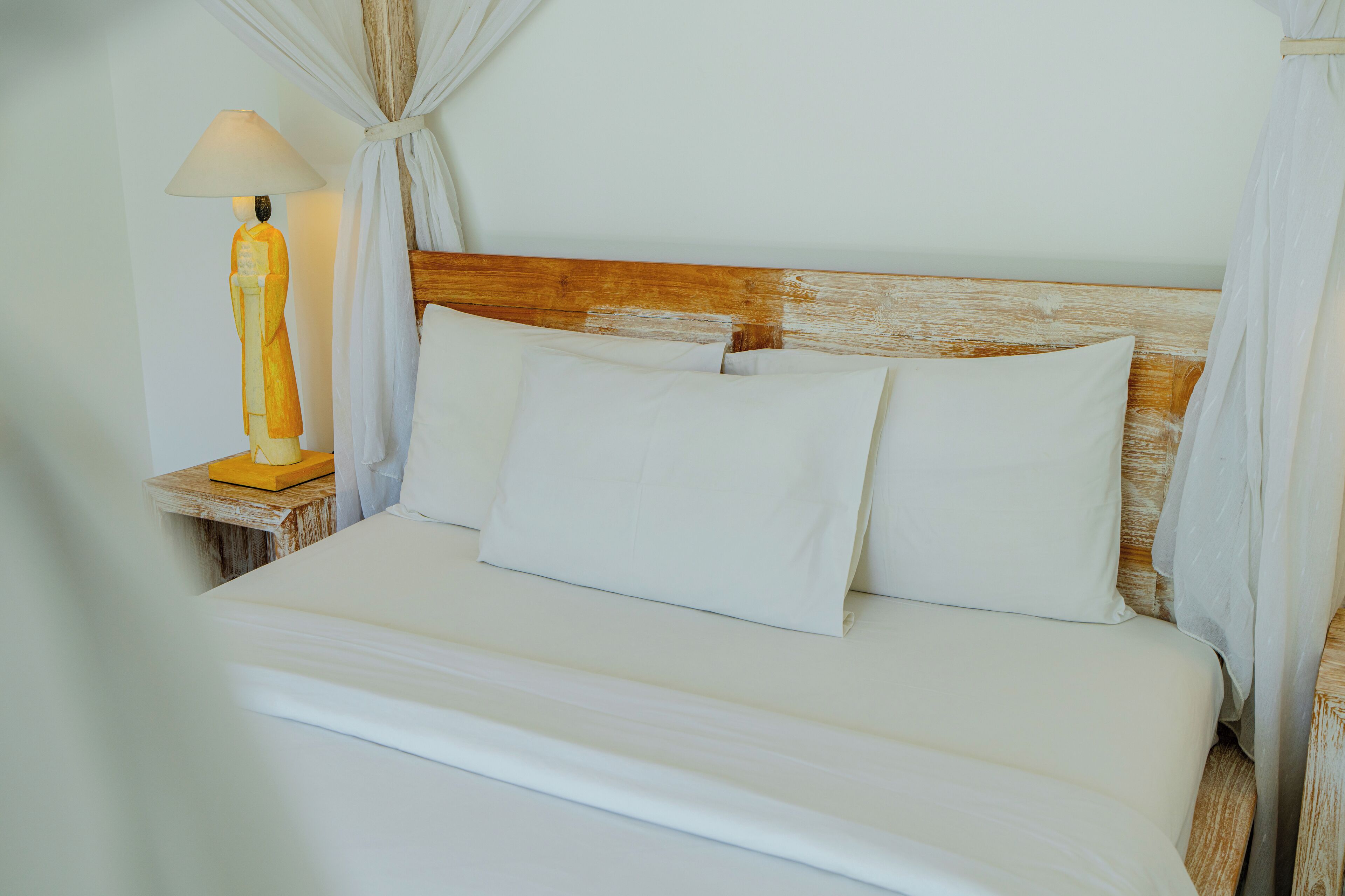 Egy gondosan megterített ágy fehér ágyneművel és párnákkal, egy fafejjel, és egy éjjeliszekrény, rajta egy lámpával.