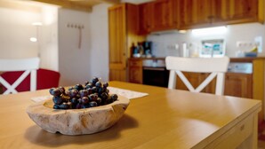 Une table en bois, sur laquelle se trouve un saladier de raisins, et une chaise blanche, dans une cuisine dotée de meubles en bois et d’un plan de travail blanc.