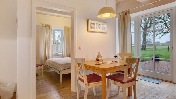 A room with a wooden dining table, four chairs, a bed, a window with a view of a green landscape, and a pendant light.