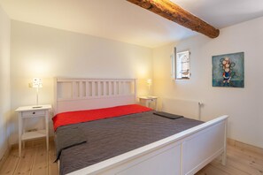 A bedroom with a white bed, red and gray bedding, two bedside tables, a window with a view, and a painting on the wall.