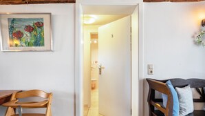 Una habitación con una silla de madera, una mesita pequeña, un cuadro enmarco, una puerta que da al baño y una cama con un almohadón.
