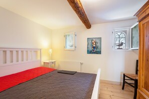 A bedroom with a bed, a red blanket, a nightstand, a lamp, a window, and a painting.