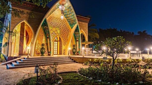 A building with a large arched entrance, steps leading up to it, and a garden area with plants and a small tree. - Shilp Wellness Resort &  Spa (Panvel)
