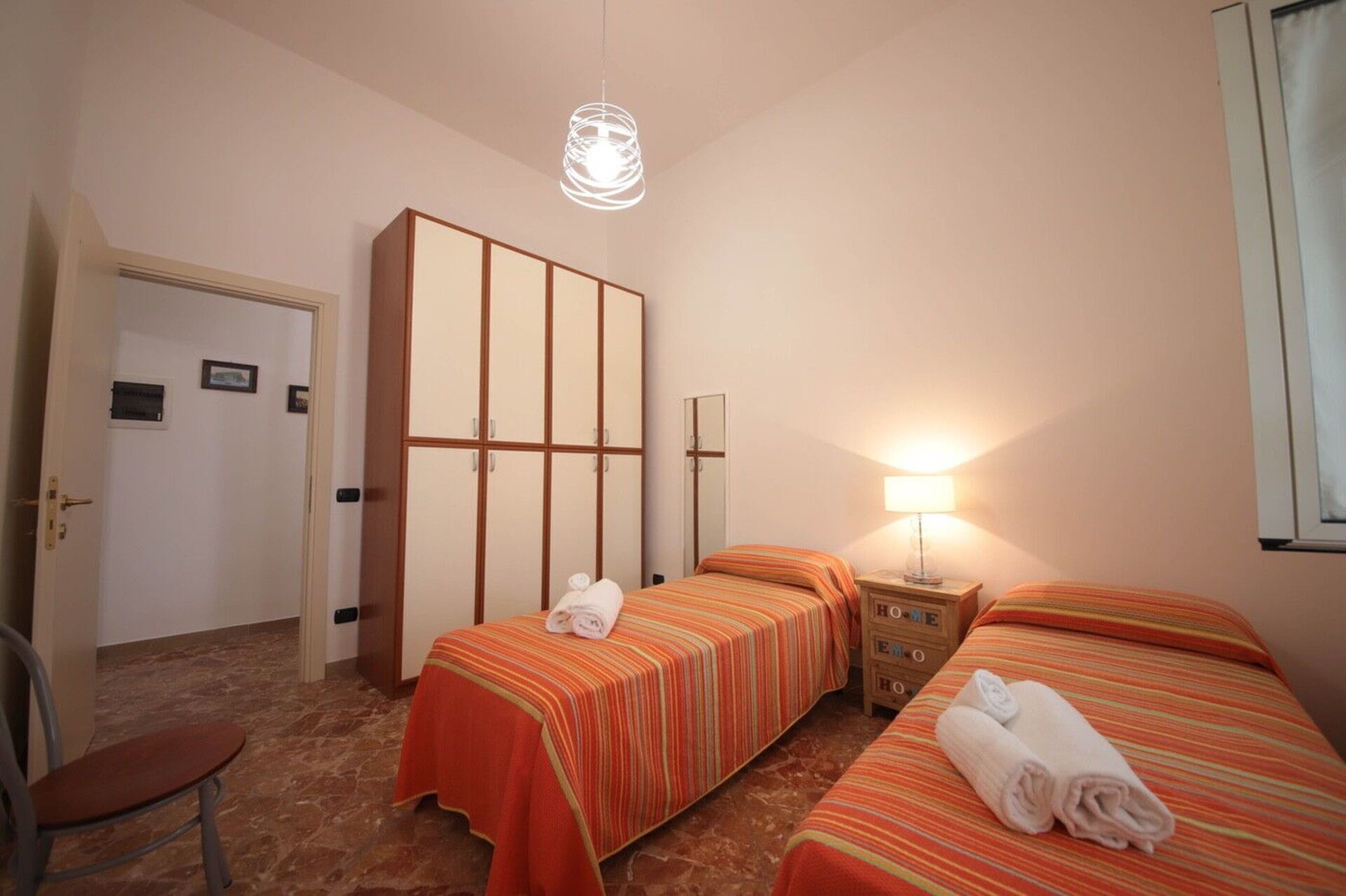 A hotel room with two beds, a chair, a wardrobe, and a lamp.