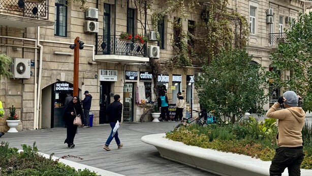 Una plaza pública con un edificio que tiene varios balcones, unidades de aire acondicionado y una variedad de tiendas, incluyendo una cafetería y un mercado.