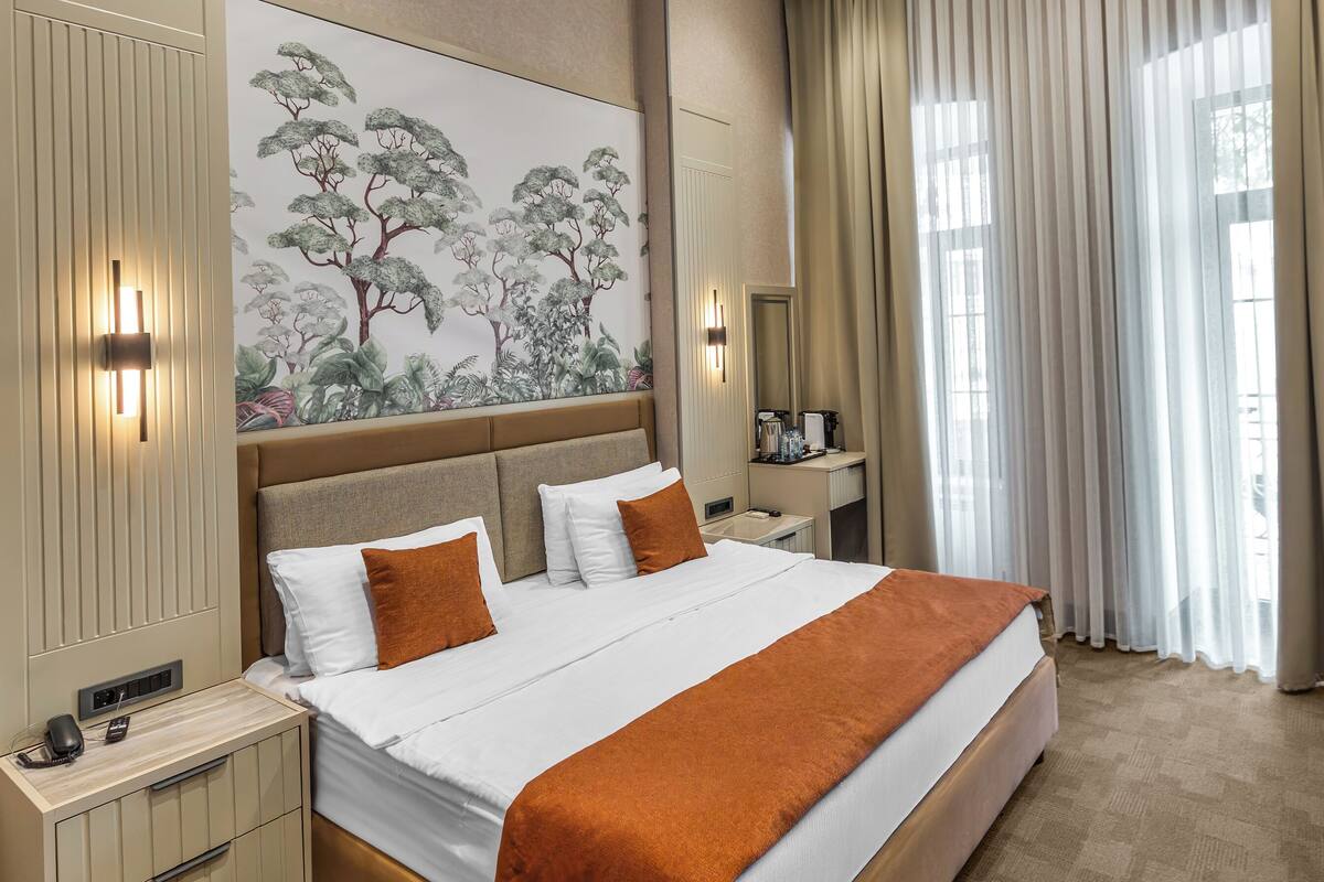Una habitación de hotel moderna con una cama grande, una mesita de noche, un escritorio y un ventanal con cortinas.