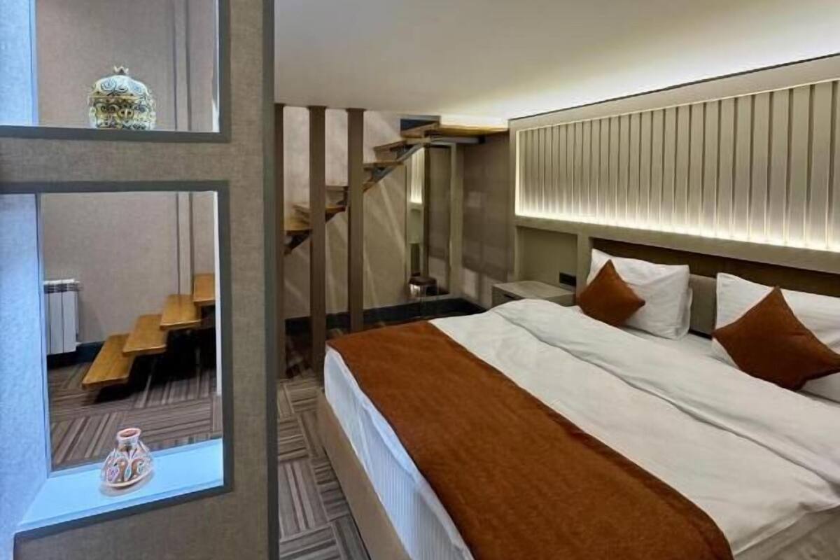 Una habitación de hotel moderna con una cama grande, cabecera con iluminación integrada, un espejo y vistas a una escalera.