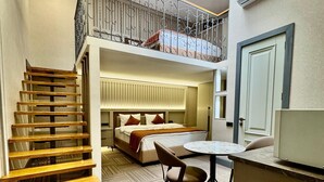 Habitación de hotel con escalera de madera, una cama con cabecera, una mesa redonda con una silla, un pequeño balcón con barandilla y un equipo de aire acondicionado en la pared.