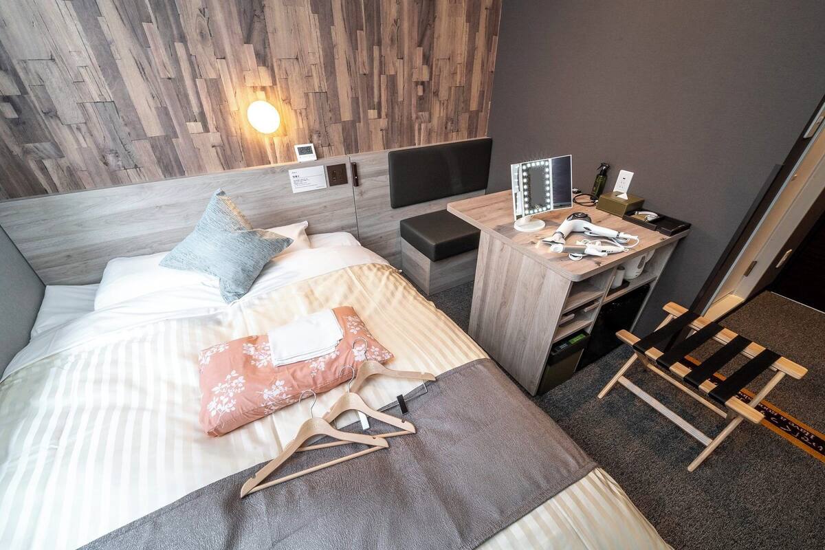 Habitació d'hotel moderna amb un capçal de llit de fusta, un llit amb roba de llit a ratlles, una tauleta de nit de fusta amb un mirall i articles de tocador, i una cadira de fusta.