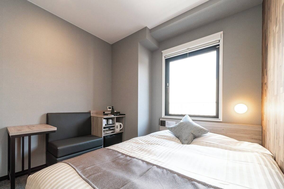 Habitació d'hotel amb un llit, una taula de nit, una cadira, una finestra amb persianes i una làmpada penjada a la paret.